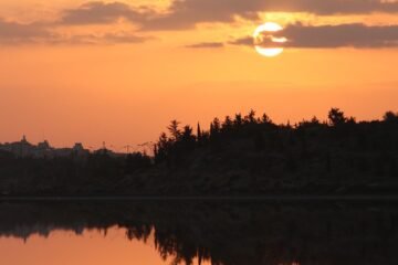 Un soare roșu care apune și se reflectă în lacul din mijlocul unei păduri de brazi